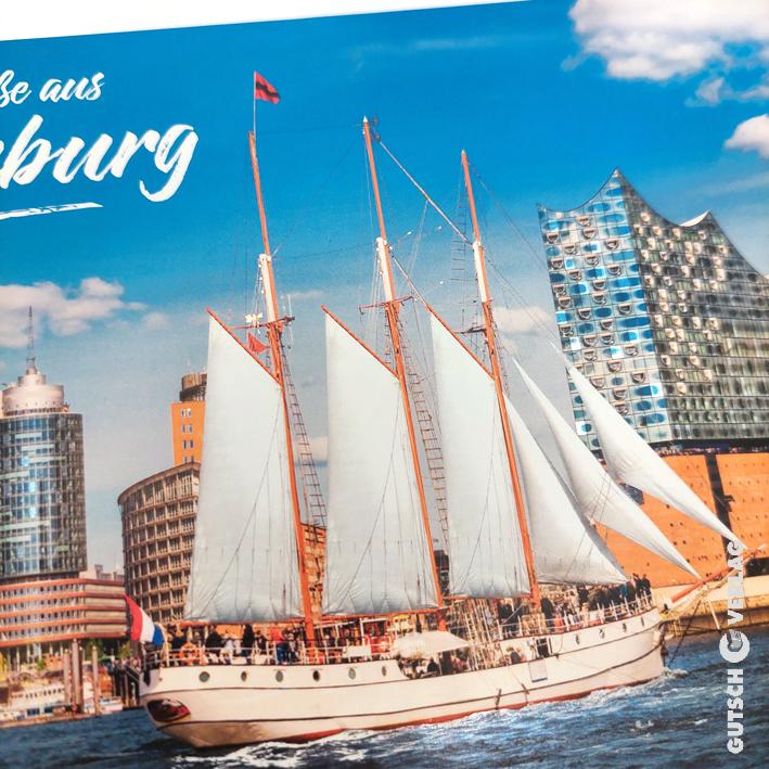 Ein historisches Segelschiff mit weißen Segeln liegt in Hamburg, im Hintergrund moderne Gebäude.