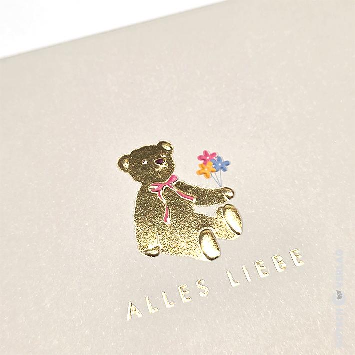 Ein Teddybär aus Goldfolie mit einer rosa Schleife und bunten Blumen auf einer strukturierten cremefarbenen Karte mit dem Text ALLES LIEBE.