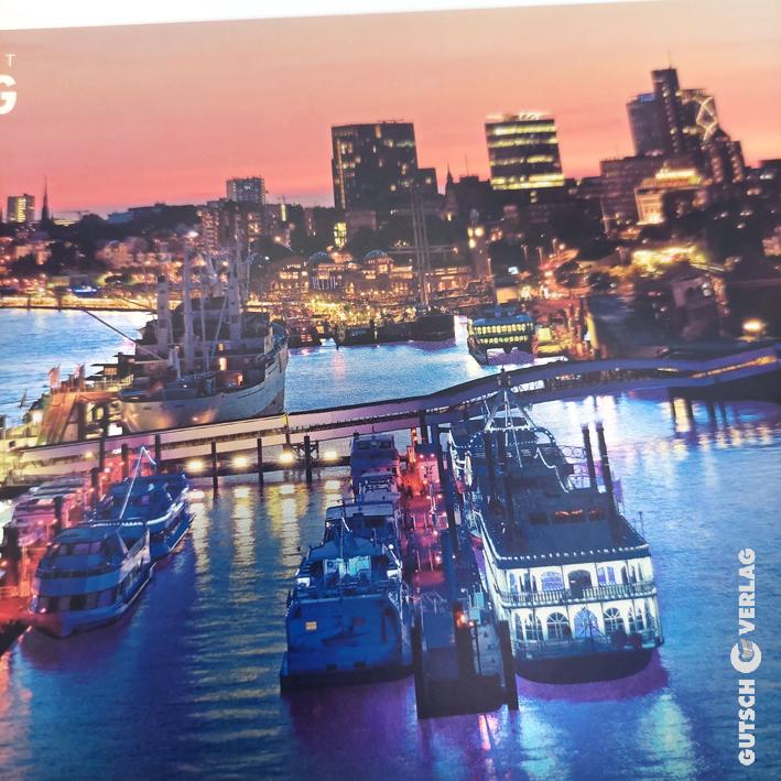 Eine lebendige Hafenszene in der Abenddämmerung mit beleuchteten Booten und der Skyline der Stadt, die sich im Wasser spiegelt.