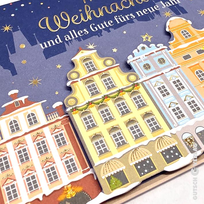 Eine festliche Weihnachtskarte mit farbenfrohen, illustrierten Gebäuden im europäischen Stil, verziert mit Goldfolienakzenten und Schnee.