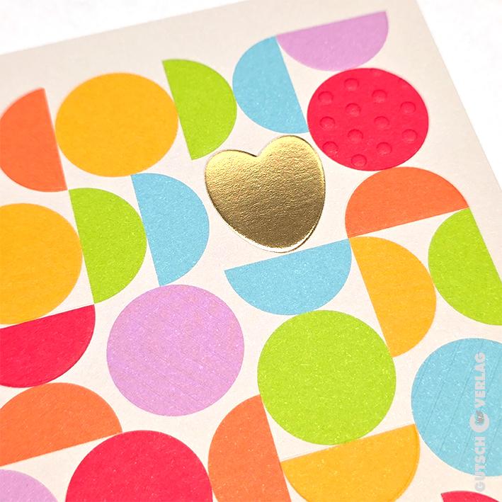 Eine Grußkarte zeigt ein Muster aus bunten Kreisen und Halbkreisen mit einer zentralen, glänzenden Goldfolien-Herzform.
