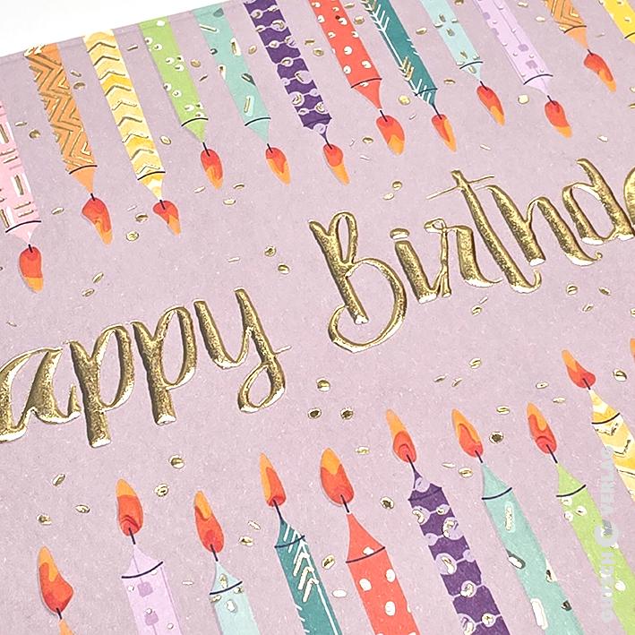 Eine lavendelfarbene Geburtstagskarte mit Reihen bunt gemusterter Kerzen und den Worten „Happy Birthday“ in Goldfolie.