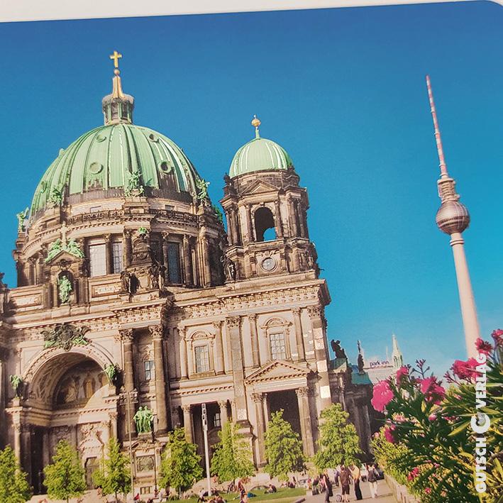 Berliner Dom mit grünen Kuppeln und Berliner Fernsehturm vor strahlend blauem Himmel, umgeben von viel Grün.