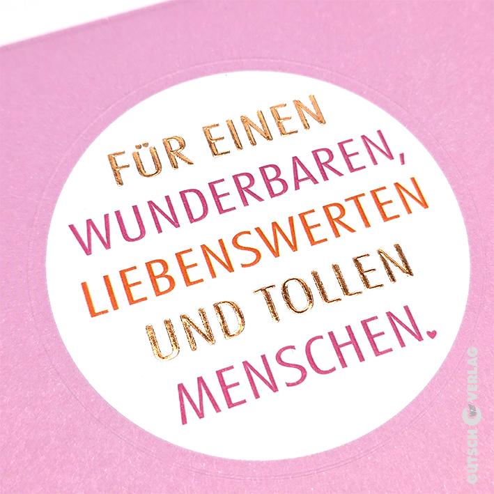 Ein runder Aufkleber auf rosa Hintergrund mit deutschem Text in Metallic-Gold und rosa Folie drückt herzliche Wünsche aus.